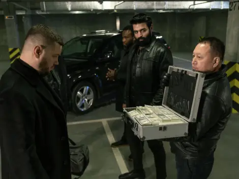 Mature man in black jacket, gloves and jeans opening suitcase with stacks of dollars while showing them to killer